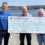 Three people stand outdoors by the sea, holding a large ceremonial check for £25,000 made out to Viking Genes from the North Yell Development Council.