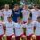 A soccer team of fourteen players, wearing white and red uniforms, poses together outdoors on grass for a group photo.