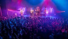 A band performs on stage under colorful lights while a large crowd watches and dances in front of the stage at an indoor concert venue.