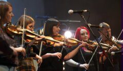 Five musicians stand on stage playing violins, with microphones and stage lights visible in the background.