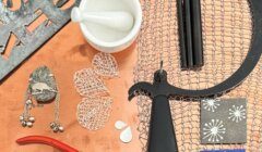 A variety of jewelry-making tools and materials, including wire mesh, pliers, stencils, a mortar and pestle, and metal components, are arranged on a work surface.