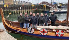 Eight men stand on a wooden Viking-style boat docked at a harbor, with shields lining the boat’s side and marine vessels visible in the background.