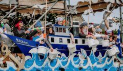 People dressed in sailor and pirate costumes sit on a parade float shaped like a ship, holding buckets and water guns, with decorations of waves and seagulls.