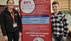 Two people stand beside a banner about young sudden cardiac death, featuring statistics and information on screening and symptoms, with contact details for the CRY screening program.