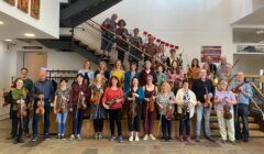 A group of people holding string instruments pose for a photo in front of a staircase indoors, with some standing on the stairs and others on the ground floor.