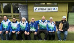 Seven people, mostly in matching blue and green jackets, sit in front of a building with signs for Lerwick Outdoor Bowling Club and Lerwick Distillery. One holds a large brown paper bag.