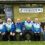 Seven people, mostly in matching blue and green jackets, sit in front of a building with signs for Lerwick Outdoor Bowling Club and Lerwick Distillery. One holds a large brown paper bag.