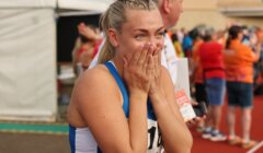 A female athlete in a blue and white uniform stands on a track, covering her mouth with both hands and appearing emotional. People and event tents are visible in the background.