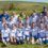 A women’s soccer team and coaches pose for a group photo on grass, wearing matching white and blue uniforms, with fields and hills in the background.