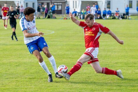 Two soccer players compete for the ball; one in a white and blue kit, the other in red, as other players and spectators watch from the background on a grassy field.