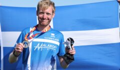 A man in a blue Team Shetland shirt holds a medal and mascot, standing in front of a large Shetland flag on a sunny day.