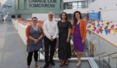 Four adults are standing side by side indoors in front of colorful bunting and a sign that reads “Change Our Tomorrow.”.
