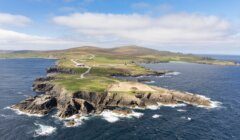 Aerial view of a rocky coastline with cliffs, green fields, scattered buildings, and winding roads surrounded by the sea under a partly cloudy sky.