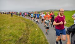 A large group of people of various ages running outdoors on a paved path beside a grassy field under an overcast sky.
