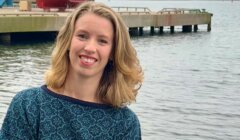 A woman with shoulder-length blonde hair stands by a waterside dock, smiling, with boats and hills visible in the background.