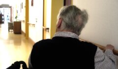 An elderly person in a wheelchair holds a handrail while sitting in a hallway with soft lighting and pale walls.