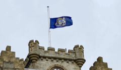 A blue flag with a seal flies at half-mast atop a stone building with castle-like features, under an overcast sky.
