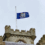 A blue flag with a seal flies at half-mast atop a stone building with castle-like features, under an overcast sky.
