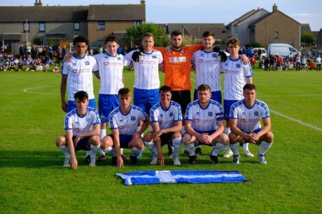 A men's soccer team in white and blue uniforms poses for a group photo on a grassy field with a flag laid out in front of them. Houses and spectators are visible in the background.