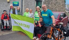 A group of people, including a child and a person in a wheelchair, stand outdoors holding a large Samaritans flag beside a bicycle and a red car.