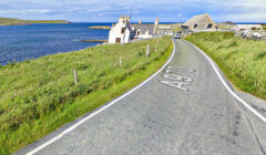 A narrow road marked A970 curves beside the sea, bordered by grassy slopes, with a few stone buildings near the shoreline under a partly cloudy sky.