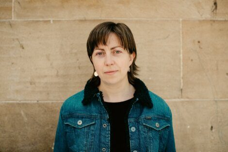 A person with short brown hair wearing a denim jacket stands in front of a beige stone wall, looking at the camera with a neutral expression.
