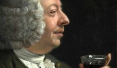 A man with white curly hair and a green coat holds a glass of dark liquid, looking at it with a slight smile.