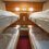 A small room with two sets of bunk beds, white linens, wood-paneled walls, and a nightstand in the middle under bright overhead lights.