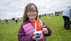 A young girl with a butterfly painted on her face holds a container of Angelito ice cream while standing on a grassy field at an outdoor event.