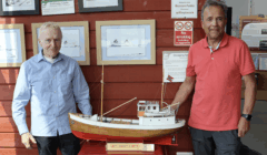 Two men stand beside a detailed model boat displayed indoors, with informational signs and framed photos on the red wall behind them.