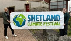 Two people stand outside holding a banner that reads "Shetland Climate Festival" with an earth graphic on it.
