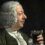 A man with white curly hair and 18th-century clothing holds a glass of dark beverage, looking at it with a slight smile against a dark background.
