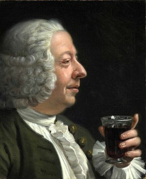 A man with white curly hair and 18th-century clothing holds a glass of dark beverage, looking at it with a slight smile against a dark background.
