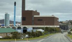 A large industrial building with a tall smokestack stands beside a road, with smaller structures and trees in the foreground under a cloudy sky.