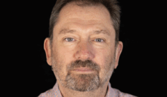 Middle-aged man with short brown hair and a beard, wearing a pink checkered shirt, facing forward against a plain black background.