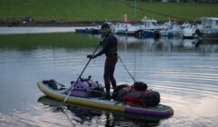 A person paddles a stand-up paddleboard loaded with bags and gear on a calm body of water near a marina with boats and grassy hills in the background.
