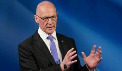A bald man in a suit and tie gestures with his hands while speaking at a podium against a blue background.