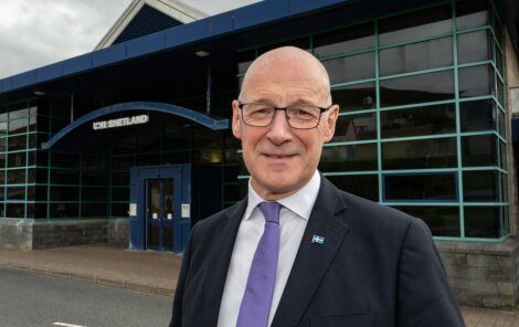 Bald man in a suit and glasses stands outside a modern building with a sign reading "UHI Shetland.