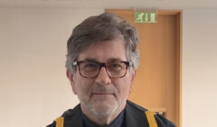 An older man with gray hair and glasses wearing a black robe with yellow trim stands indoors near a wooden door and an emergency exit sign.