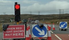A red traffic light and road signs indicate construction ahead, with cones and vehicles blocking part of a rural road under cloudy skies.