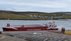 A red cargo ship named "BALTIC WAVE" is docked at a port, carrying a large wind turbine blade on its deck. Hilly terrain and industrial buildings are visible in the background.