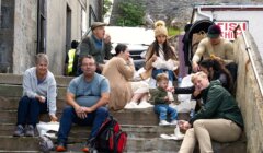 A group of people, including adults and children, sit on outdoor stone steps eating food, with bags and containers around them.