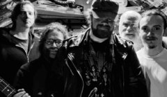 Five men stand together in a dimly lit room filled with posters and memorabilia; the central figure wears a leather jacket and cap. The image is in black and white.