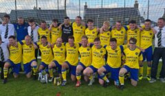 A men's soccer team in yellow and blue uniforms poses together in front of a goal, with coaches in dress shirts beside them. Two trophies are displayed on the grass in front.