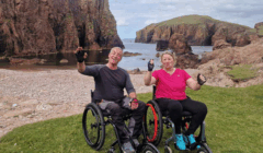 Two people in wheelchairs sit on grass by a rocky coastline, smiling and waving at the camera with cliffs and the sea in the background under a cloudy sky.