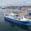 A large blue and white fishing vessel docked at an industrial port with other ships and buildings visible in the background.