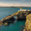 A lighthouse stands on a rocky promontory with a natural stone arch over the sea, surrounded by blue water and distant hills under a partly cloudy sky.
