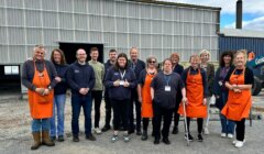 A group of people, some wearing orange aprons and name badges, pose together outdoors in front of a metal industrial building.