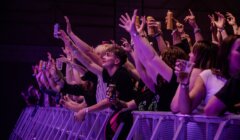 A crowd of people at a concert stands behind a barricade, holding drinks and reaching toward the stage under purple lighting.