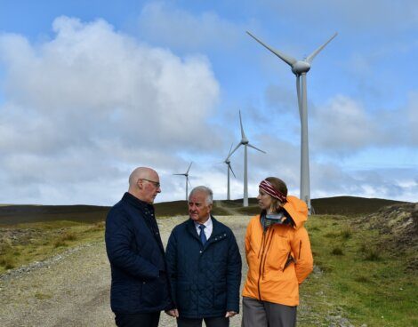 Three people stand on a dirt path in front of wind turbines on a grassy landscape, having a conversation under a partly cloudy sky.